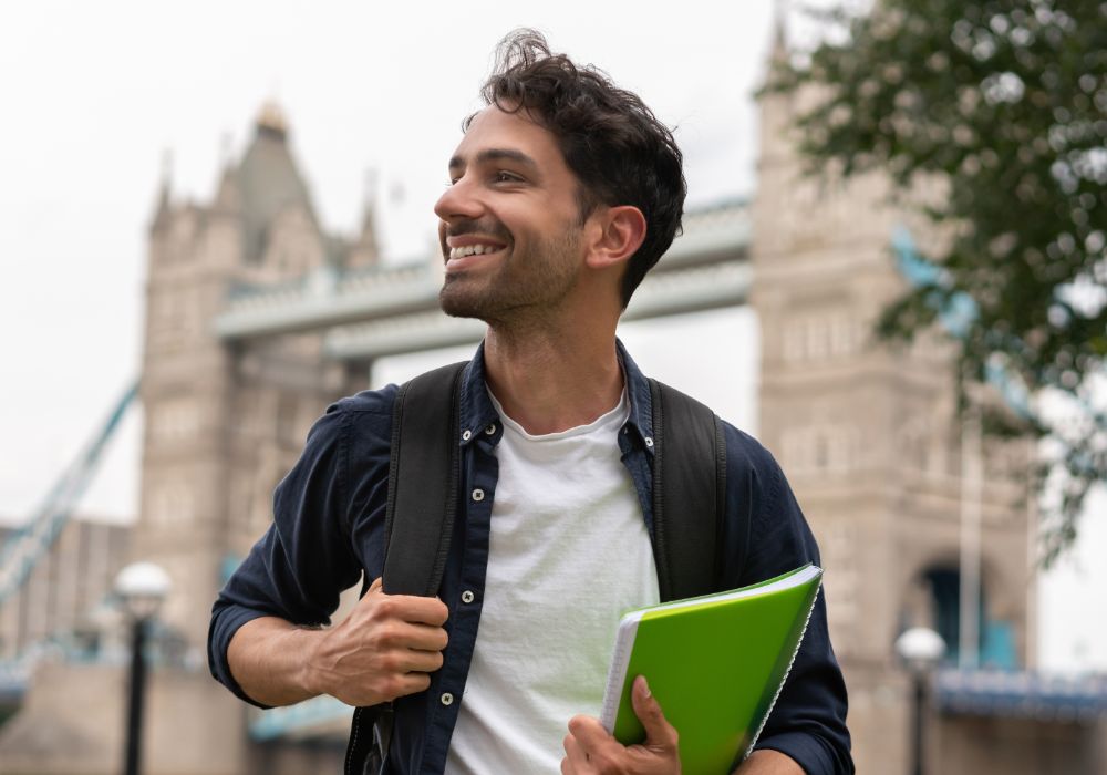 A man with a backpack stands in front of Tower Bridge.